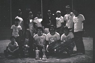 YFC baseball team poses with championship trophy, 1970s. Indoor stadium setting with visible net and lighting. Uniforms featu...
