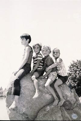 Four children pose playfully on a large rock by water, mid-20th century. Boys in striped swim trunks and tank tops, girls in ...
