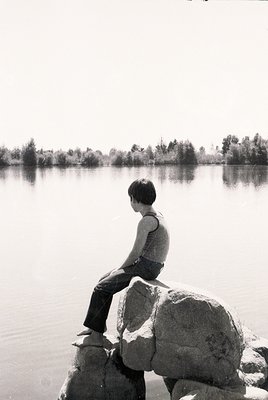A solitary figure sits on jagged rocks by a serene lake, gazing across calm waters. Mid-century streetwear—sleeveless shirt, ...