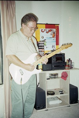 Mid-century indoor setting featuring a man playing a sunburst-toned electric guitar, likely a Fender-style model. Surrounding...