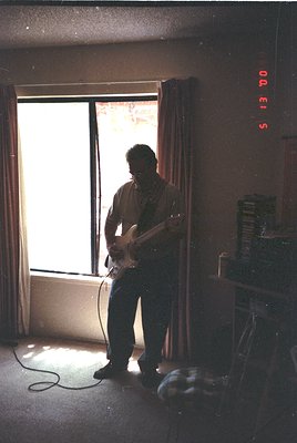 Mid-century guitar studio with analog clock (5:13 PM) showing time. Man in casual 1960s–70s attire plays electric guitar near...