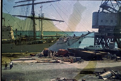 Vintage sepia-toned shipyard scene with a multi-masted sailing vessel docked alongside industrial cranes and scaffolding. For...