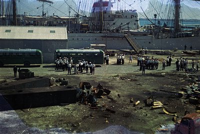 Vintage port scene with industrial docks, mid-20th century. Workers in uniform stand near debris and cargo, with a large ship...