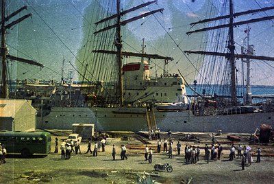 Vintage sepia-toned maritime scene featuring a large four-masted sailing ship docked at a port, surrounded by smaller vessels...