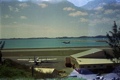 Vintage aerial view of a coastal airfield with twin-engine propeller aircraft on a grass runway. Mid-20th century hangar and ...