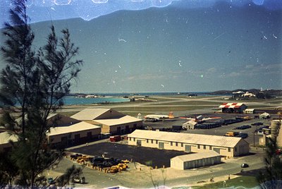 Vintage aerial view of a coastal airfield with mid-century concrete buildings, parked aircraft, and vehicles. Mountain range ...