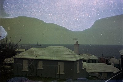 Vintage coastal scene through aged glass—distorted but revealing: low-slung beach houses with flat roofs, a chimney, and util...