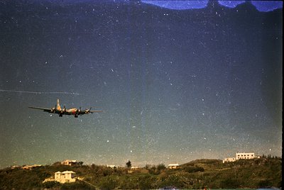 A vintage propeller aircraft ascends at night, illuminated by distant city lights against a star-studded sky. Likely a 1950s–...