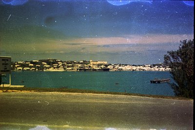 Vintage seaside town with low-rise buildings along a calm coastal road, captured through a slightly yellowed filter. Dense fo...