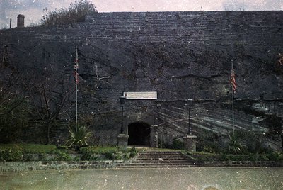 Historic stone entrance tunnel with arched doorway, flanked by two flagpoles. Surrounding vegetation includes sparse trees an...