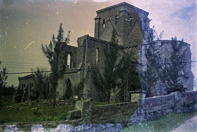 Vintage sepia-toned photo of a ruined Gothic-style stone church with arched windows and a bell tower, partially overgrown wit...