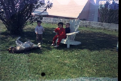 Vintage snapshot of three children in a backyard setting, likely mid-20th century. Two boys sit on a white Adirondack chair, ...