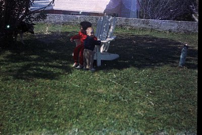 Two children in vintage clothing (1960s-70s) play on wet grass beside a metal bench. One wears a red coat, the other a stripe...