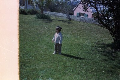 A child in mid-20th century attire stands on a well-manicured lawn, framed by a window edge. Lighting suggests vintage color ...