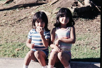 Two young girls in 1970s-style striped tank tops and shorts sit on a concrete ledge, eating. Grass and rocks in the backgroun...
