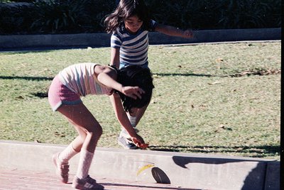 Two children mid-air on a concrete surface, likely a playground, mid-1970s. Girl in striped shirt and shorts leaps forward, b...