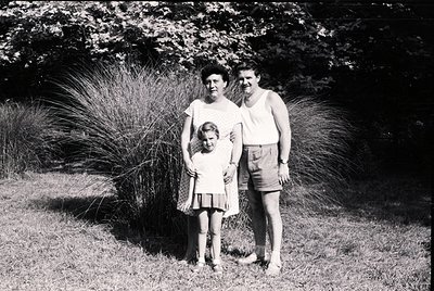 Family portrait in mid-20th century outdoor setting, likely 1950s–1960s. Three individuals pose on grass beside ornamental gr...