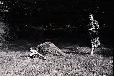 Mid-20th century rural scene: Woman in floral dress (1950s-60s style) stands beside haystack, while child lies on grass. Lush...