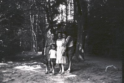 Three women and a child pose outdoors in a wooded area, likely mid-20th century. The woman on the left wears a patterned dres...