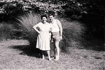 Couple posing outdoors in mid-20th century attire—woman in a knee-length floral dress, man in sleeveless shirt and shorts. Lu...