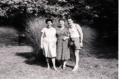 Three individuals pose outdoors in mid-20th-century attire, likely 1950s–1960s. Woman on left wears a sleeveless dress with a...