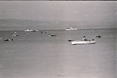 Mid-20th century seaside scene: crowded beach with swimmers, a lone rowboat, and distant boats. Worn grainy texture suggests ...