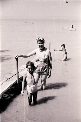 A woman in a floral swimsuit and headband leads a young boy in swim diapers along a shallow beach edge, 1950s-60s. Another ch...