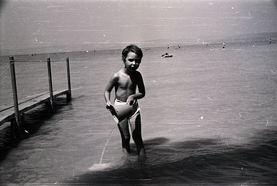 Black-and-white shot of a young boy in mid-20th-century swim trunks, standing in shallow water near a wooden pier. His focuse...