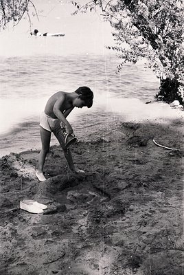 Mid-20th-century beachgoer in shorts and sandals kneels to place a small wooden box on sandy shore, near shallow water. Class...