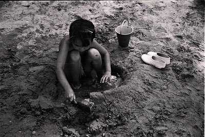 Child kneeling in muddy excavation, holding small shovel. Nearby: bucket and worn sandal. Mid-20th century agricultural or co...