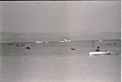 Black-and-white coastal scene featuring mid-20th century beachgoers in shallow waters. Small wooden rowboat and scattered swi...