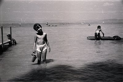 Mid-20th century seaside scene: shirtless boy in swim trunks holds a bucket near a wooden pier, wading into shallow water. Ot...