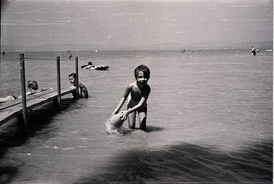 Mid-20th century beach scene with wooden pier and shallow waters. A young boy in swim trunks holds a bucket, wading near the ...