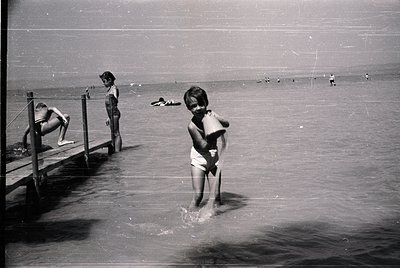 Mid-20th century seaside scene: children wade in shallow water near a wooden pier. Foreground boy in swim trunks clutches a t...