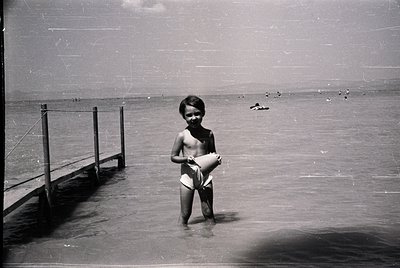 Young boy in mid-20th-century swim trunks stands waist-deep in shallow coastal waters, clutching a white cloth or towel. Wood...