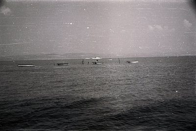 Vintage black-and-white seascape showing three small wooden boats anchored near shore, likely fishing vessels. Distant shorel...