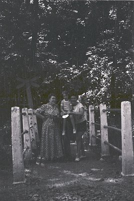 Three individuals pose on a rustic wooden bridge with a cross in a forested area, mid-20th century. The woman wears a pattern...