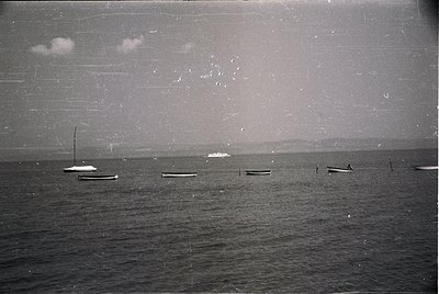 Vintage black-and-white seascape featuring small wooden fishing boats anchored in calm waters. Distant shoreline with faint i...