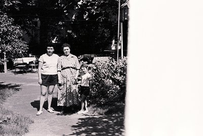 Black-and-white family portrait in a residential garden, mid-20th century. Adults in casual summer attire—man in short-sleeve...