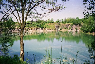 Quarry lake reflecting clear blue skies, surrounded by exposed rock cliffs and lush greenery. Overgrown tree branch frames le...