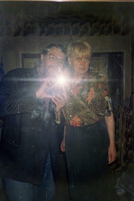 Vintage portrait of two individuals posing indoors, likely 1970s–1980s. Man in dark jacket, round glasses, and patterned tie;...