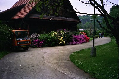 Vintage orange utility vehicle parked beside a lush garden bed bursting with vibrant pink, yellow, and purple flowers. Wooden...