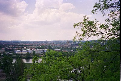 Panoramic view of a mid-20th century European cityscape with a river winding through urban and green zones. Distinctive churc...