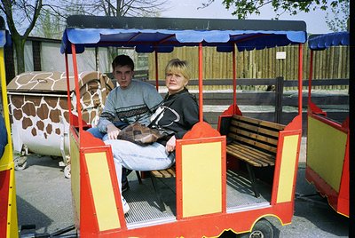 Vintage zoo tram with bright red/blue canopy carrying two passengers in 1980s-90s attire. Giraffe enclosure with patterned sk...