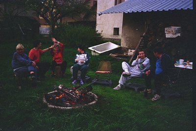 Family gathering around an outdoor fire pit in a residential courtyard, likely 1990s. Six adults and children seated on stool...