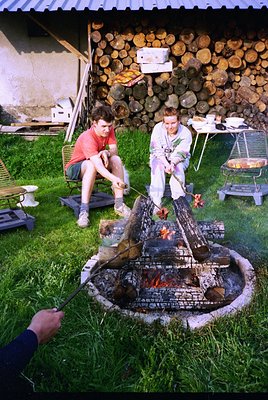 Two individuals tend a brick-built outdoor fire pit, skewering meat over flames. Stacked logs line the background wall. Mid-2...