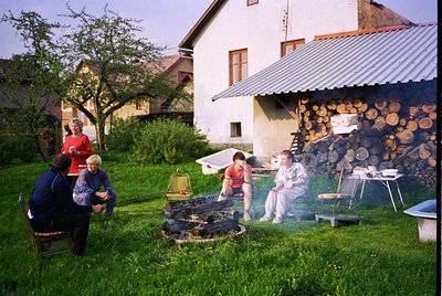 Family gathers around a rustic outdoor barbecue setup in a lush garden, with a woodpile and metal grill emitting smoke. Mid-2...