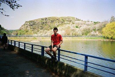 Young man in red polo shirt sits on blue-painted concrete railing beside calm water, with forested hillside and clear sky in ...