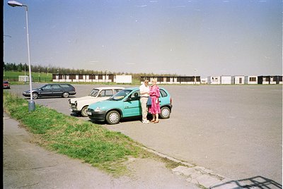 Vintage 1990s-era parking lot with two women near a turquoise hatchback. One wears a pink dress, the other a striped top. Whi...