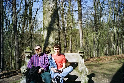 Two men pose on a weathered stone bench in a forested cemetery, surrounded by bare trees and gravestones. The man on left wea...